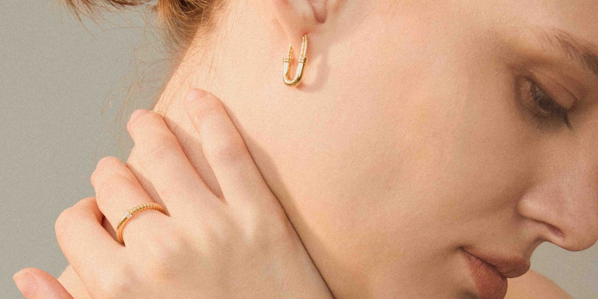 Close-up of a woman wearing a small gold hoop earring and a gold ring on her finger, with her hand resting near her neck against a soft gray background.